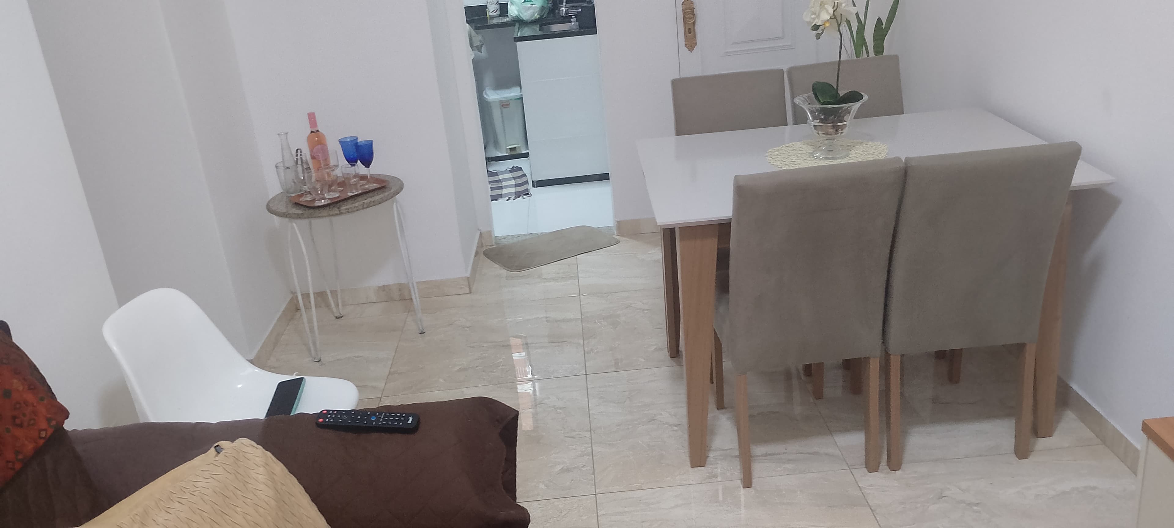 Sala de jantar com mesa branca e quatro cadeiras estofadas em tom neutro, com vista parcial para a cozinha.