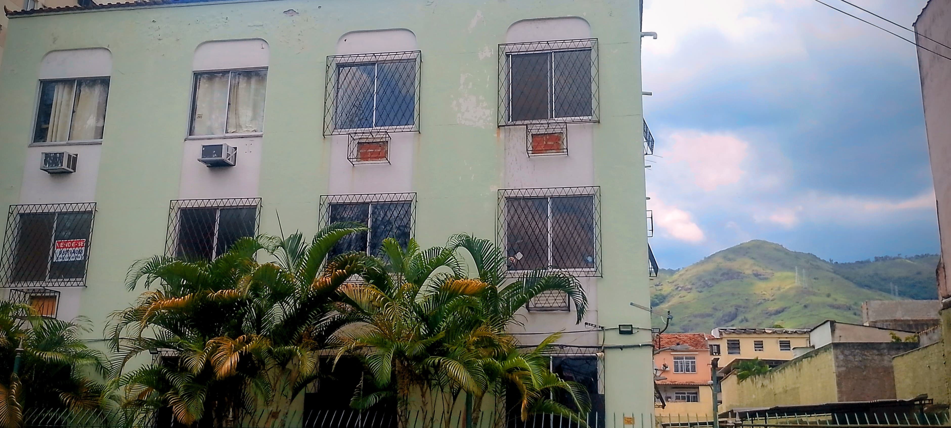Fachada de prédio residencial com pintura clara, janelas com grades e vegetação na frente.