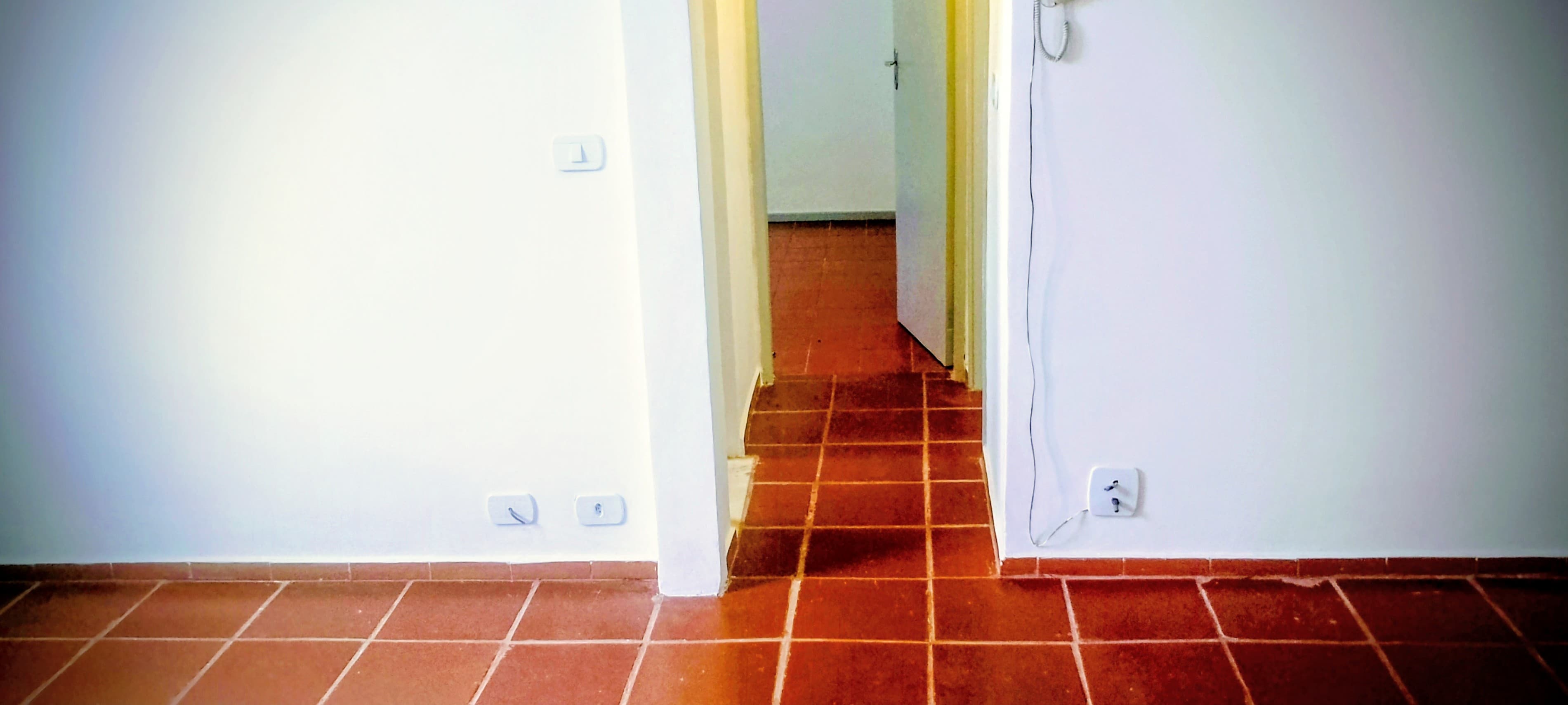 Ambiente interno com paredes brancas, piso de cerâmica avermelhada e porta de acesso a outro cômodo.